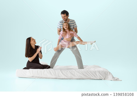 Father holding daughter in split jump above white bed while mother applauds Father holding daughter in split jump above white bed while mother applauds 139858108