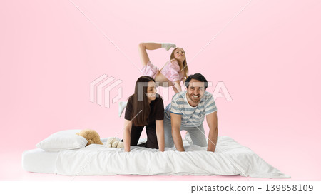 Smiling family playing on white bed with daughter climbing on parents backs. Smiling family playing on white bed with daughter climbing on parents backs. 139858109