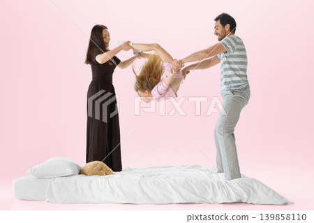 Parents swinging daughter above white bed in playful pastel pink bedroom scene. 139858110
