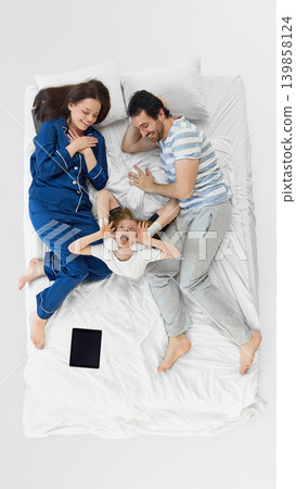 Playful daughter making funny face between parents on white bedding. 139858124