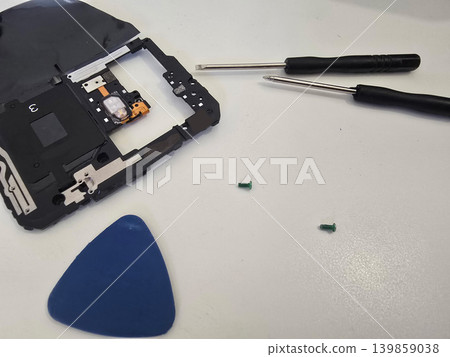 A technician repairs a modern smartphone with exposed internal components, battery, and tools on a white workbench. Concept for electronics repair and mobile device servicing A technician repairs a modern smartphone with exposed internal components, battery, and tools on a white workbench. Concept for electronics repair and mobile device servicing 139859038