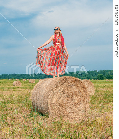 Woman standing on a large hay bale in a summer field, wearing a flowing dress and sunglasses under a bright sky Woman standing on a large hay bale in a summer field, wearing a flowing dress and sunglasses under a bright sky 139859202