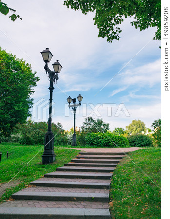 A winding stone staircase ascends through a vibrant green park, flanked by two classic black lampposts. Lush trees and bushes surround the path 139859208