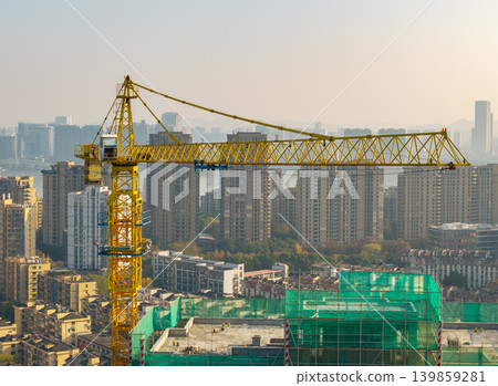 A large yellow tower crane stands prominently above a new building site covered in green netting in a modern developed city A large yellow tower crane stands prominently above a new building site covered in green netting in a modern developed city 139859281