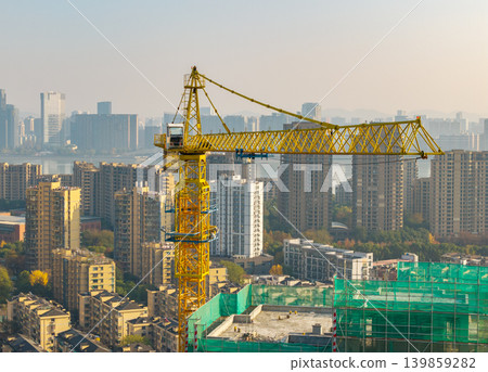 A large yellow tower crane stands prominently above a new building site covered in green netting in a modern developed city A large yellow tower crane stands prominently above a new building site covered in green netting in a modern developed city 139859282