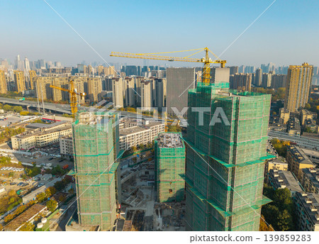 Aerial view of a bustling city showcasing ongoing construction of multiple high rise buildings covered with scaffolding netting and with large cranes operating Aerial view of a bustling city showcasing ongoing construction of multiple high rise buildings covered with scaffolding netting and with large cranes operating 139859283