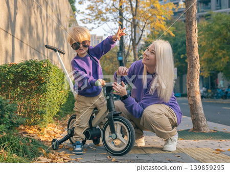Smiling mother crouches next to her young son on a balance bike on a brick sidewalk. They wear matching purple shirts during a bright autumn afternoon 139859295