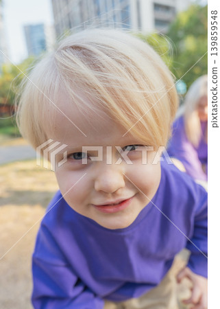 A young child with light hair enjoys time in a park wearing a purple shirt. The child looks directly at the camera with a smile A young child with light hair enjoys time in a park wearing a purple shirt. The child looks directly at the camera with a smile 139859548