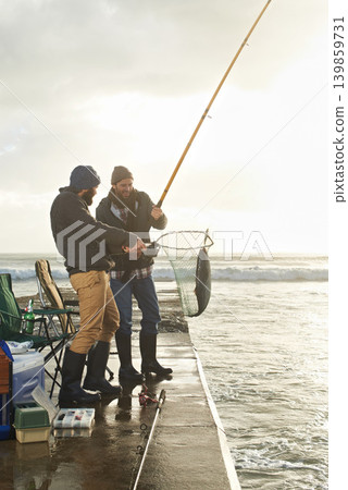 Fishing, men and net with fish at pier with rod, waves and relax on vacation, holiday and adventure. Friendship, people and bonding in morning with overcast, sky and nature for activity and hobby 139859731