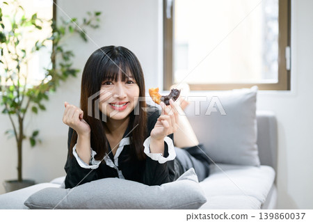 Japanese woman relaxing in bed while eating a donut. Japanese woman relaxing in bed while eating a donut. 139860037