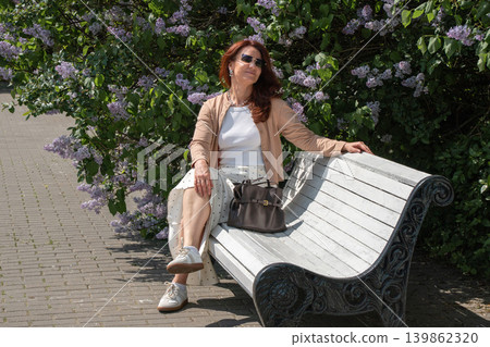 Red-haired woman in sunglasses relaxes on white park bench beside blooming lilac bushes and sunny pedestrian walkway in spring garden. Lifestyle, wellness, city leisure, urban recreation, social media Red-haired woman in sunglasses relaxes on white park bench beside blooming lilac bushes and sunny pedestrian walkway in spring garden. Lifestyle, wellness, city leisure, urban recreation, social media 139862320