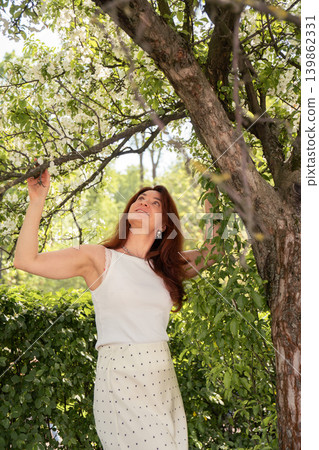 Red-haired woman in white outfit stands beneath blooming apple tree branches with soft sunlight, green foliage, spring garden atmosphere. Lifestyle, wellness branding, nature, spring fashion, outdoor Red-haired woman in white outfit stands beneath blooming apple tree branches with soft sunlight, green foliage, spring garden atmosphere. Lifestyle, wellness branding, nature, spring fashion, outdoor 139862331