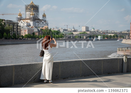 Red-haired woman photographs Cathedral of Christ the Saviour from Moscow River embankment under blue sky with city skyline, water view. Tourism, mobile photography, travel, city landmark, social media Red-haired woman photographs Cathedral of Christ the Saviour from Moscow River embankment under blue sky with city skyline, water view. Tourism, mobile photography, travel, city landmark, social media 139862371