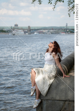 Red-haired woman relaxes on embankment in Gorky Park, Moscow River under warm summer sunlight with urban waterfront. Lifestyle, wellness branding, city leisure, travel advertising, summer recreation Red-haired woman relaxes on embankment in Gorky Park, Moscow River under warm summer sunlight with urban waterfront. Lifestyle, wellness branding, city leisure, travel advertising, summer recreation 139862372