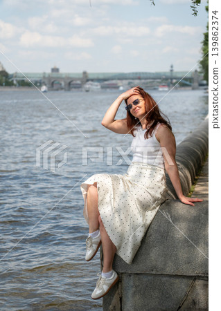 Red-haired woman in sunglasses sits on Gorky Park embankment beside Moscow River with bridge, city skyline in summer light. Lifestyle, wellness, city leisure, travel advertising, summer recreation Red-haired woman in sunglasses sits on Gorky Park embankment beside Moscow River with bridge, city skyline in summer light. Lifestyle, wellness, city leisure, travel advertising, summer recreation 139862374