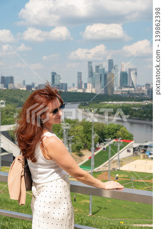 Red-haired woman overlooks Moscow skyline from observation deck near Moscow River with green parkland, modern towers under summer clouds. Tourism, city travel, lifestyle, urban panorama, social media 139862398