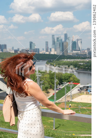 Red-haired woman enjoys panoramic view of Moscow River and Moscow City skyscrapers from observation deck under bright summer sky with soft clouds. Tourism, city travel, urban panorama, social media 139862402