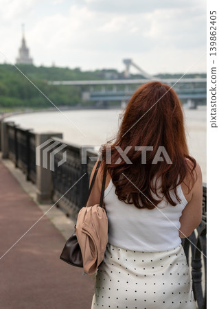 Red-haired woman looks over Moscow River from embankment with bridge, Sparrow Hills cableway and Moscow State University silhouette. Tourism, city travel, riverside leisure, social media 139862405