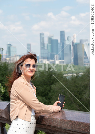 Red-haired woman with smartphone stands on Sparrow Hills observation deck with Moscow City skyline and green forest landscape. Tourism, city travel advertising, mobile technology Red-haired woman with smartphone stands on Sparrow Hills observation deck with Moscow City skyline and green forest landscape. Tourism, city travel advertising, mobile technology 139862408