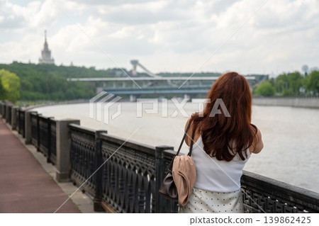 Red-haired woman looks over Moscow River from embankment with bridge, Sparrow Hills cableway and Moscow State University silhouette. Tourism, city travel, riverside leisure, social media Red-haired woman looks over Moscow River from embankment with bridge, Sparrow Hills cableway and Moscow State University silhouette. Tourism, city travel, riverside leisure, social media 139862425