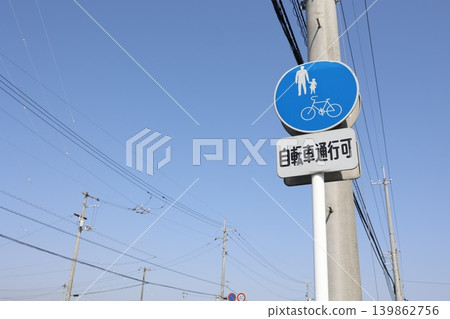 A sign indicating that bicycles are allowed on the sidewalk. A utility pole and a blue sky. A blue sign indicating that bicycles are permitted. 139862756