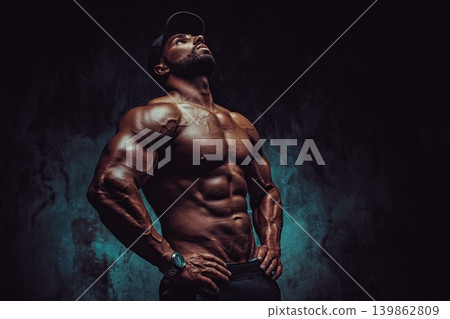 Young strong man bodybuilder in cap on wall background, dramatic colors 139862809