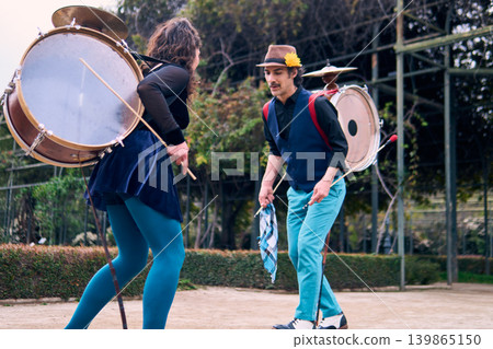 Adult couple of urban dancers, happily playing and dancing the cueca with a bass drum on their backs outdoors, Chilean chinchineros, street musicians who play the bass drum and dance in the streets, Adult couple of urban dancers, happily playing and dancing the cueca with a bass drum on their backs outdoors, Chilean chinchineros, street musicians who play the bass drum and dance in the streets, 139865150