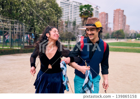 Adult couple of urban dancers, happily playing and dancing the cueca with a bass drum on their backs outdoors, Chilean chinchineros, street musicians who play the bass drum and dance in the streets. 139865158