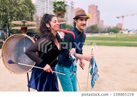 Adult couple of urban dancers, happily playing and dancing the cueca with a bass drum on their backs outdoors, Chilean chinchineros, street musicians who play the bass drum and dance in the streets. Adult couple of urban dancers, happily playing and dancing the cueca with a bass drum on their backs outdoors, Chilean chinchineros, street musicians who play the bass drum and dance in the streets. 139865159