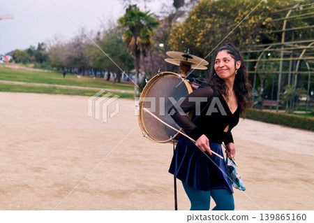 Beautiful Chilean Latina woman smiling confidently and dancing with her drum and cymbals on her back. A percussionist, street musician, dancing and playing her drum in the streets. Chilean heritage Beautiful Chilean Latina woman smiling confidently and dancing with her drum and cymbals on her back. A percussionist, street musician, dancing and playing her drum in the streets. Chilean heritage 139865160
