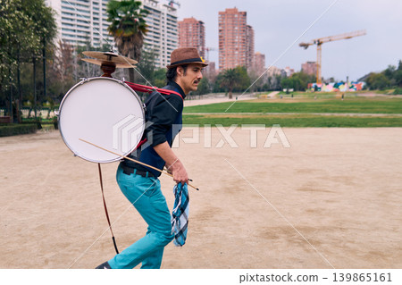 Adult Latino man, dressed in formal and colorful attire, playing a bass drum on his back outdoors, Chilean chinchinero, street musician playing the bass drum and dancing in the streets 139865161
