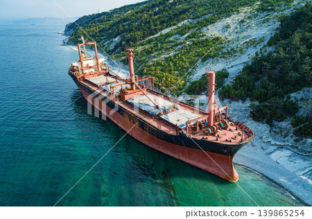 Stranded bulk carrier resting on a rocky shore beside a vibrant green forested hillside during daytime Stranded bulk carrier resting on a rocky shore beside a vibrant green forested hillside during daytime 139865254