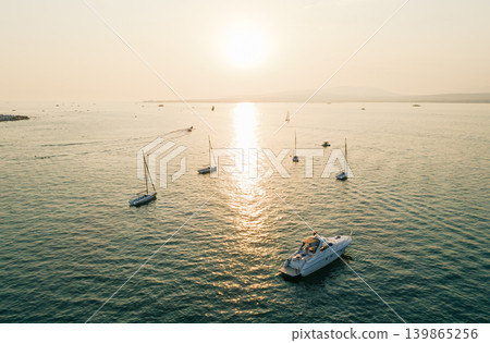 Aerial view of numerous sailing boats and a large motor yacht spread across the water during a vibrant golden sunset Aerial view of numerous sailing boats and a large motor yacht spread across the water during a vibrant golden sunset 139865256