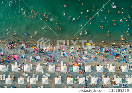 Numerous people enjoy a summer day at the beach. Swimmers cool off in the turquoise sea while others sunbathe under rows of white umbrellas and lounge chairs Numerous people enjoy a summer day at the beach. Swimmers cool off in the turquoise sea while others sunbathe under rows of white umbrellas and lounge chairs 139865259