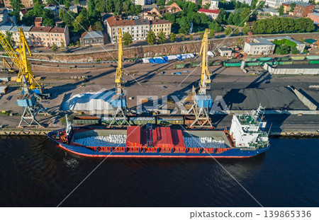 Aerial scene of an active industrial port. Yellow cranes load bulk materials into cargo ships docked at the waterfront Aerial scene of an active industrial port. Yellow cranes load bulk materials into cargo ships docked at the waterfront 139865336
