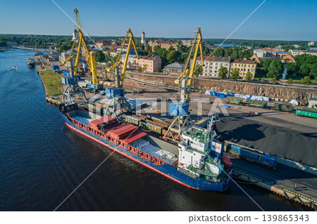 Aerial view of a blue bulk carrier ship docked at a port. Yellow cranes move materials between train wagons and the vessel 139865343