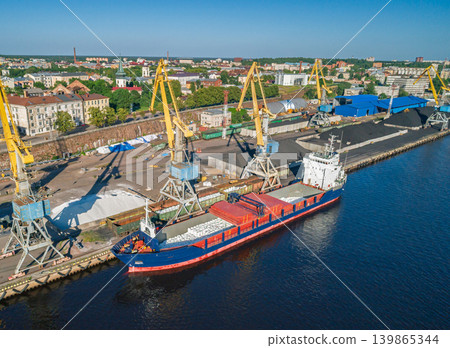 Aerial scene of an active industrial port. Yellow cranes load bulk materials into cargo ships docked at the waterfront Aerial scene of an active industrial port. Yellow cranes load bulk materials into cargo ships docked at the waterfront 139865344