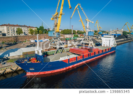 Aerial scene of an active industrial port. Yellow cranes load bulk materials into cargo ships docked at the waterfront Aerial scene of an active industrial port. Yellow cranes load bulk materials into cargo ships docked at the waterfront 139865345