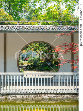 Circular opening in a white wall frames a vibrant Chinese garden scene with lush green trees and a striking red maple Circular opening in a white wall frames a vibrant Chinese garden scene with lush green trees and a striking red maple 139865395