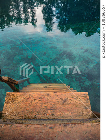 A set of wooden stairs leads into clear turquoise water. Reflections of trees and sky are visible on the water's surface, obscuring the bottom of the spring. 139866657