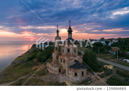 An old, crumbling Russian Orthodox church with two domes stands on a grassy cliff above a wide river during sunset An old, crumbling Russian Orthodox church with two domes stands on a grassy cliff above a wide river during sunset 139866669