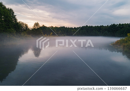 Serene morning mist covers peaceful lake surrounded by green forest. Serene morning mist covers peaceful lake surrounded by green forest. 139866687