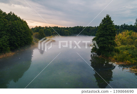 Serene morning mist covers peaceful lake surrounded by green forest. Serene morning mist covers peaceful lake surrounded by green forest. 139866690