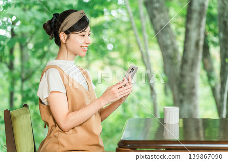 一位女顧客正在一家可以俯瞰鬱鬱蔥蔥森林的咖啡館裡使用智慧型手機。 139867090