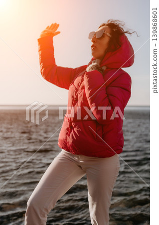 Woman sunset jacket standing by the sea in winter clothing while shielding her eyes from the sun Woman sunset jacket standing by the sea in winter clothing while shielding her eyes from the sun 139868601
