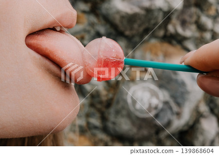 Lollipop tongue candy young woman licking a round red sweet held by another person against a rock wall Lollipop tongue candy young woman licking a round red sweet held by another person against a rock wall 139868604