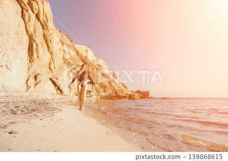 Woman beach swimsuit walks along a sandy coastline beneath high cliffs during a warm sunset by the sea Woman beach swimsuit walks along a sandy coastline beneath high cliffs during a warm sunset by the sea 139868615
