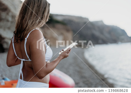 Woman smartphone beach. Young woman relaxing on a pebble beach using her phone by cliffs. 139868628