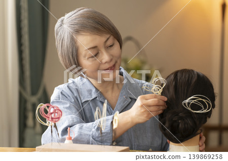 一位老婦人用水引繩製作髮飾，並將其插入人體模型中。 139869258