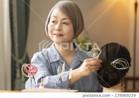 一位老婦人用水引繩製作髮飾，並將其插入人體模型中。 139869259